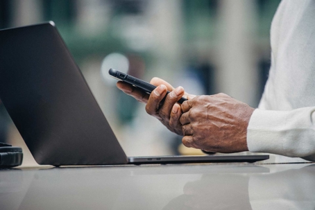 A close shot depicts a dark-skinned individual using their smartphone next to a laptop.