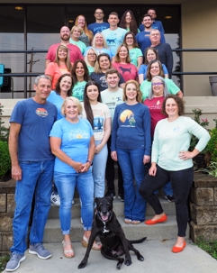 Group photo of smiling people, in casual wear, posing outside on steps, and a large black dog sits in front.
